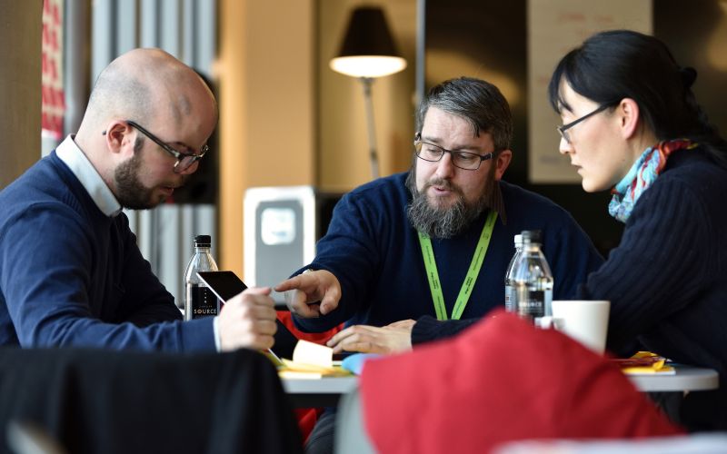 A group of people working on a group project, seated around a table looking at a laptop.