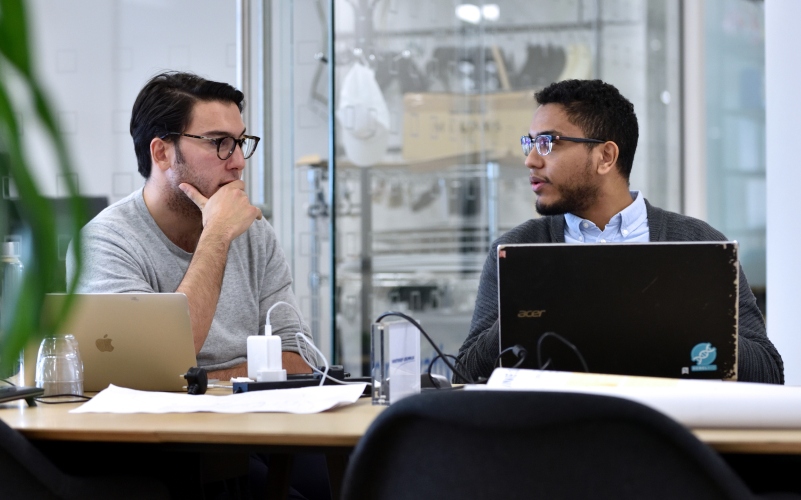 Two people working on a group project, seated at a table each with a laptop.