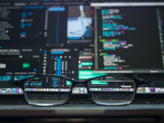 A blurred computer screen with a pair of focused glasses stting on a desk in front.